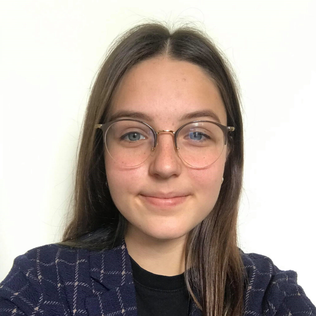 Young person with long brown hair and glasses, wearing a dark checkered blazer and black shirt, smiling softly in front of a plain white background.