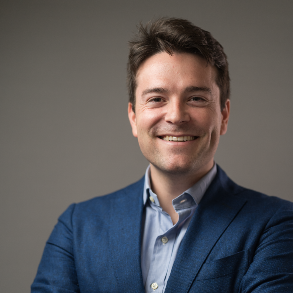 A man in a blue blazer and light blue shirt smiles with arms crossed, standing against a plain gray background.