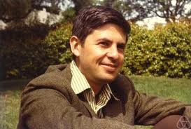 A man with dark hair, wearing a brown jacket and striped shirt, sits on grass outside, smiling, with trees and bushes in the background.