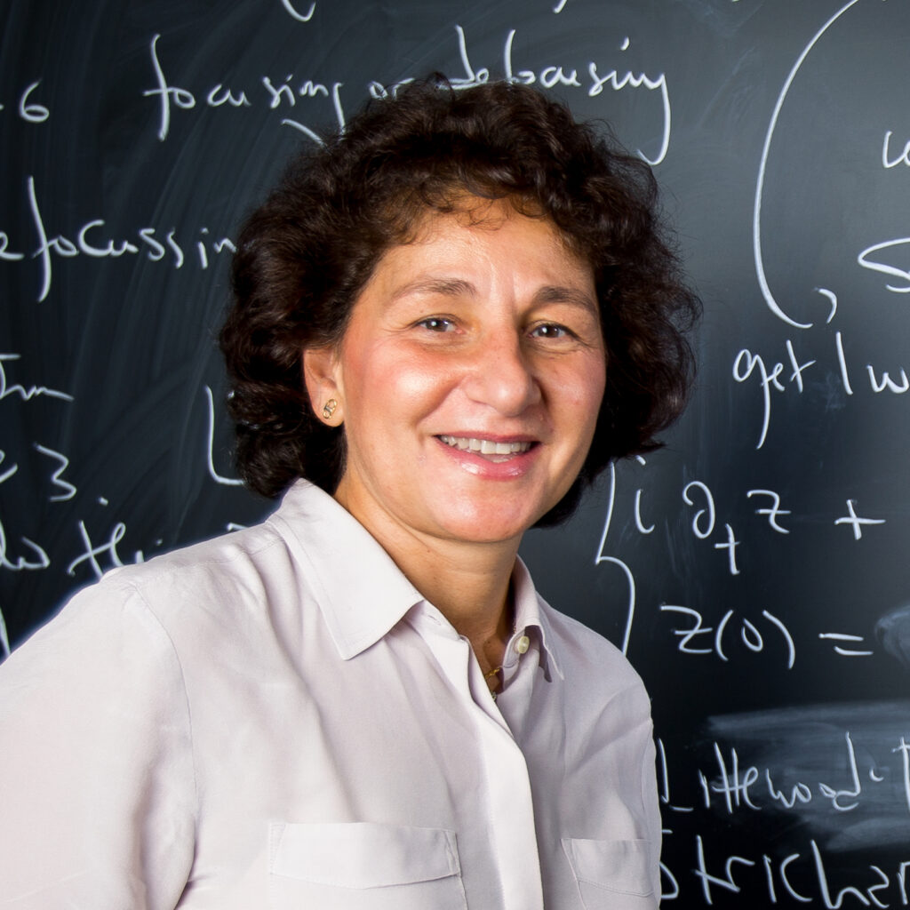 A woman with curly brown hair wearing a light-colored blouse smiles in front of a blackboard covered in mathematical equations written in white chalk.