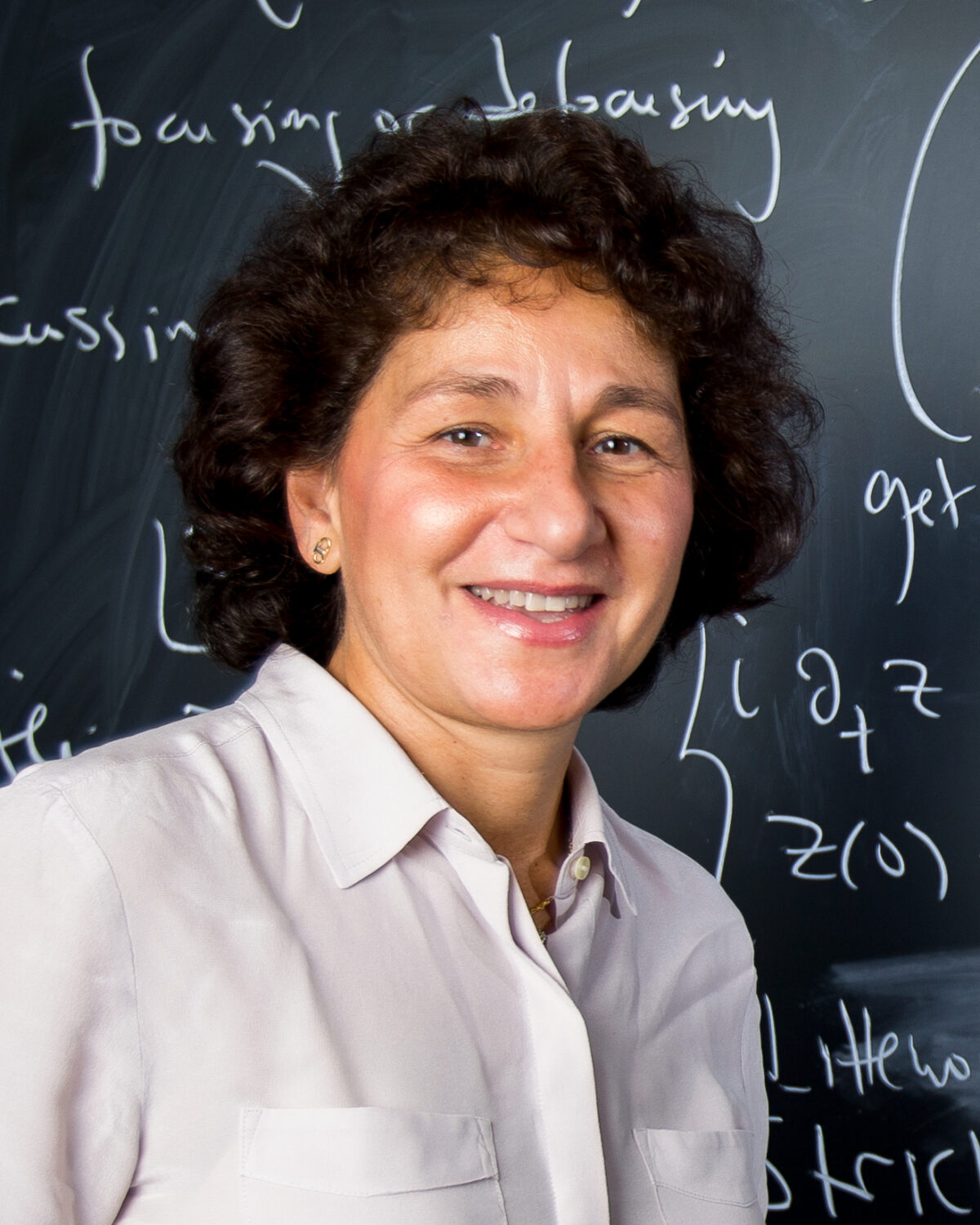 A woman with curly brown hair wearing a light-colored blouse smiles in front of a blackboard covered in mathematical equations written in white chalk.