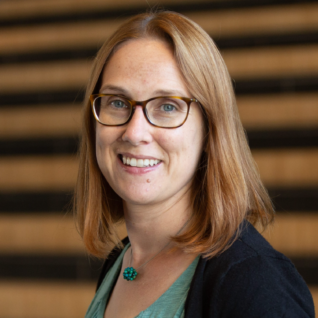 A woman with shoulder-length light brown hair and glasses smiles at the camera. She is wearing a green top, a black cardigan, and a turquoise necklace. The background features blurred horizontal lines.