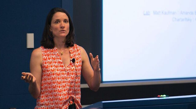 A woman with shoulder-length brown hair, wearing a sleeveless orange patterned blouse, speaks and gestures with her hands while standing in front of a presentation screen.