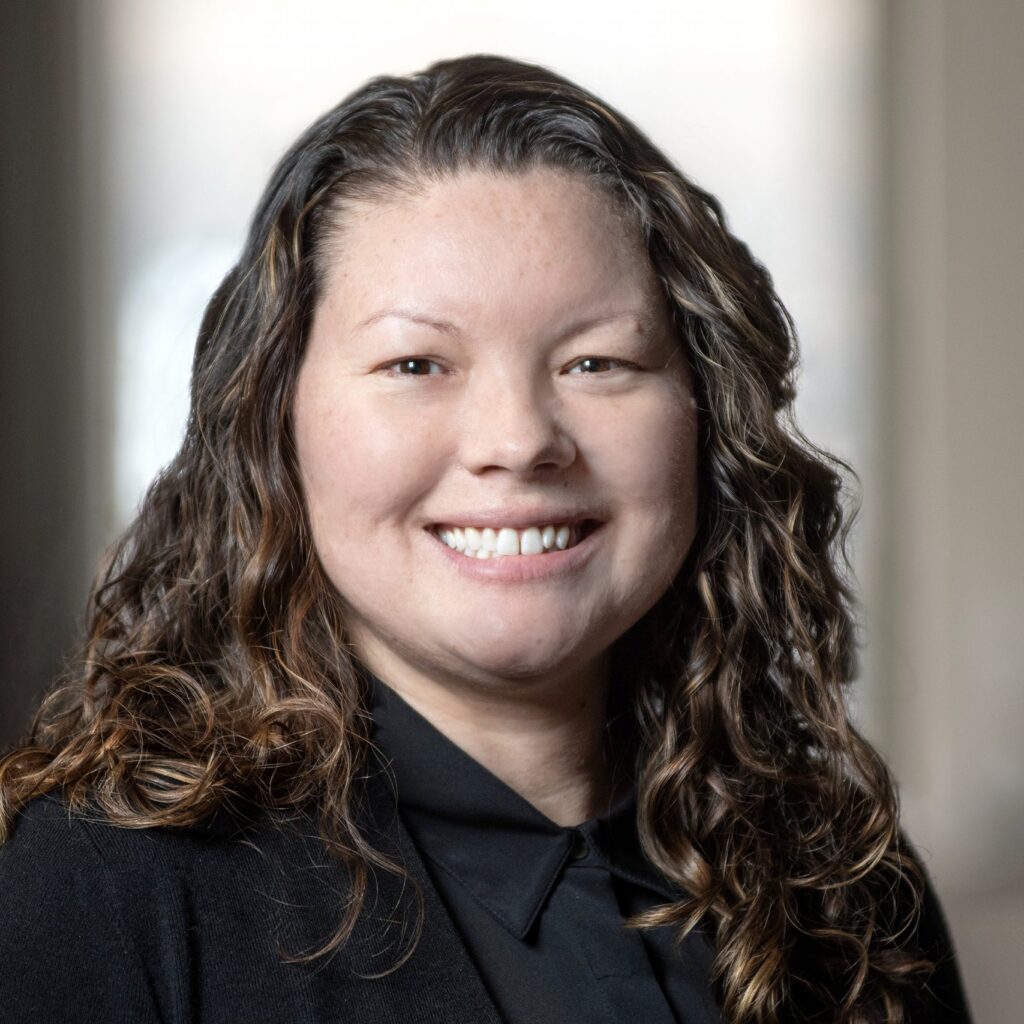 A woman with long curly brown hair, wearing a black blazer and black shirt, smiles at the camera in a softly lit indoor setting.