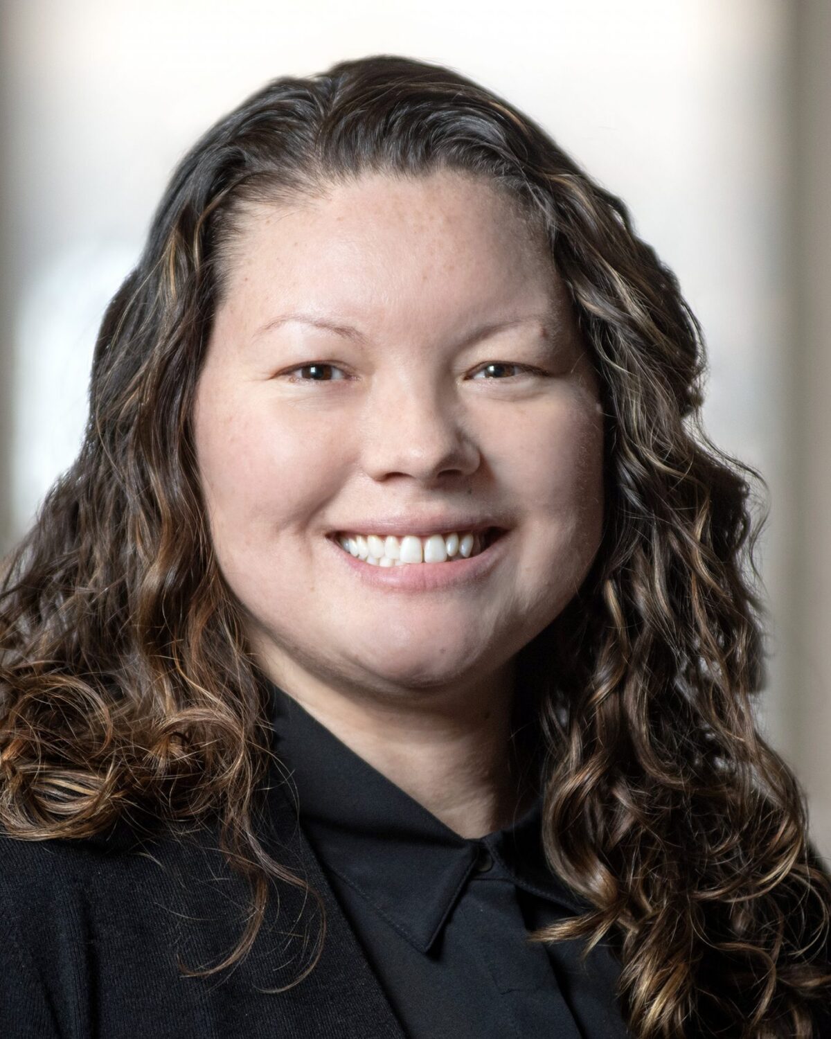 A woman with long curly brown hair, wearing a black blazer and black shirt, smiles at the camera in a softly lit indoor setting.