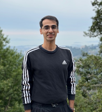 A person wearing glasses and a black Adidas sweatshirt stands outdoors, smiling, with trees and a distant, hazy landscape in the background.