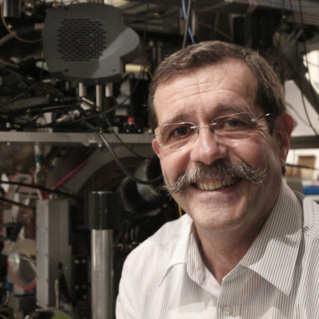 A smiling man with glasses and a prominent mustache stands in front of scientific equipment and machinery, wearing a white striped shirt.