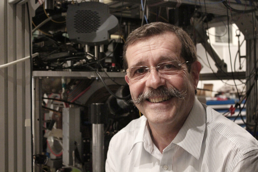 A smiling man with glasses and a prominent mustache stands in front of scientific equipment and machinery, wearing a white striped shirt.