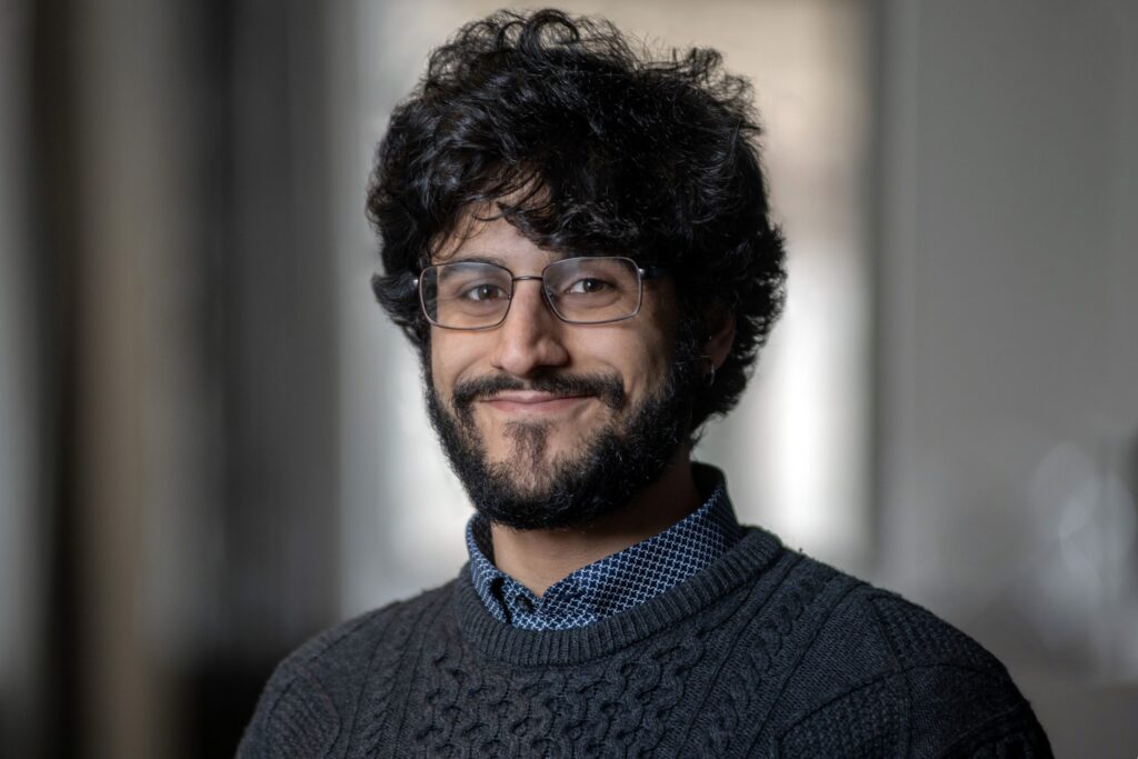 A person with curly dark hair, a beard, and glasses smiles softly, wearing a dark knit sweater and a patterned collared shirt, with a blurred indoor background.