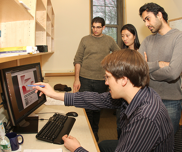 Four people in an office gather around a computer. One person is seated, pointing at a graph on the monitor, while the others stand closely, watching and listening attentively. Shelves and a window are in the background.