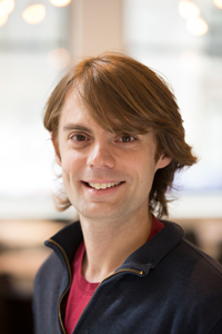 A smiling person with light brown hair wearing a dark zip-up jacket and red shirt stands indoors with soft, blurred lights in the background.