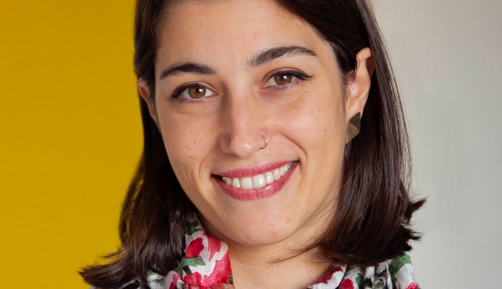 Woman with straight dark hair and a nose ring, smiling at the camera. She is wearing a colorful scarf and standing in front of a yellow and white background.