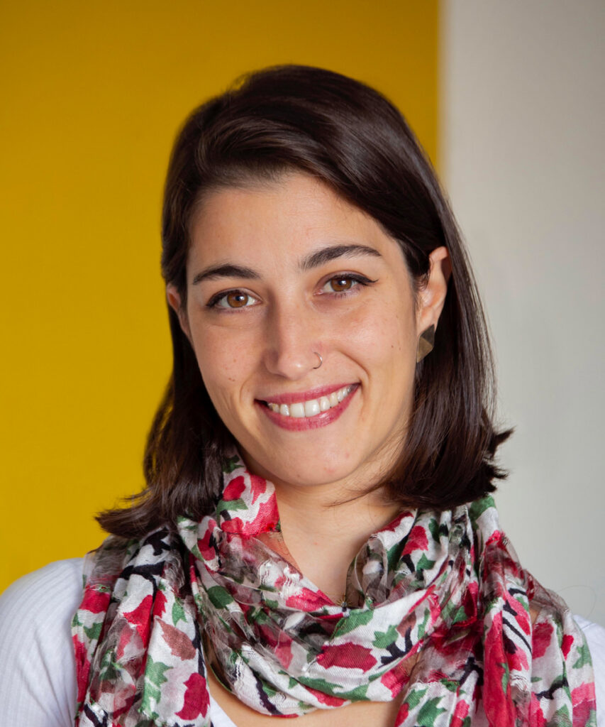 Woman with straight dark hair and a nose ring, smiling at the camera. She is wearing a colorful scarf and standing in front of a yellow and white background.