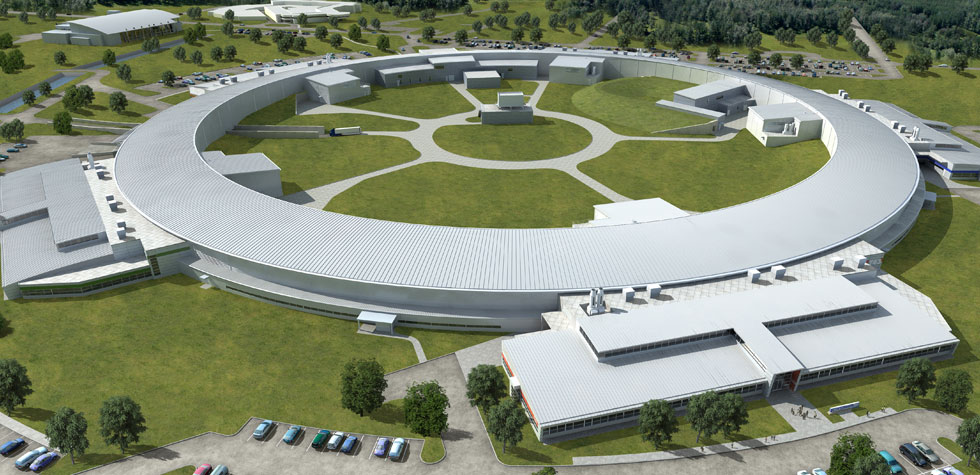 Aerial view of a large circular scientific research facility surrounded by green lawns, roads, parked cars, and several smaller buildings.