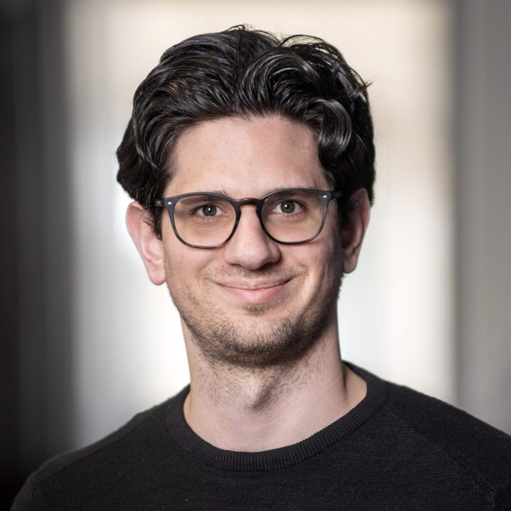 A man with wavy dark hair, glasses, and a black sweater smiles at the camera. The background is softly blurred in neutral tones.
