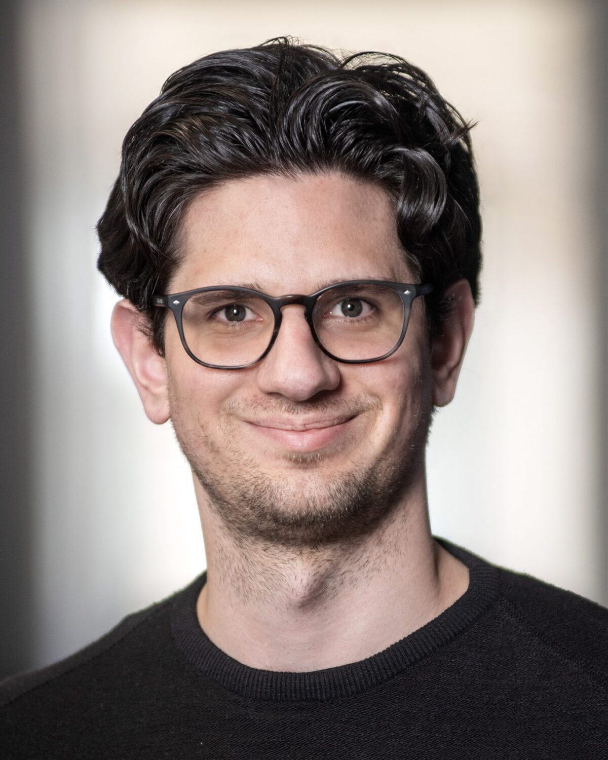 A man with wavy dark hair, glasses, and a black sweater smiles at the camera. The background is softly blurred in neutral tones.