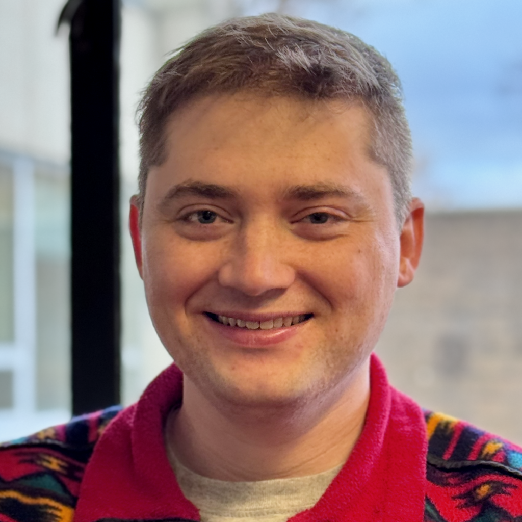 A person with short light brown hair smiles at the camera, wearing a colorful patterned sweater. The background is out of focus, showing a window and an outdoor scene.