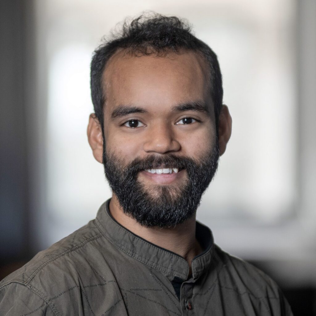 A man with a dark beard and short curly hair smiles at the camera, wearing a buttoned-up shirt. The background is softly blurred, giving a professional portrait feel.