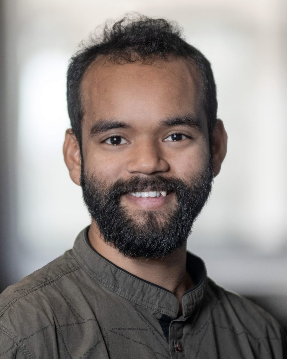 A man with a dark beard and short curly hair smiles at the camera, wearing a buttoned-up shirt. The background is softly blurred, giving a professional portrait feel.