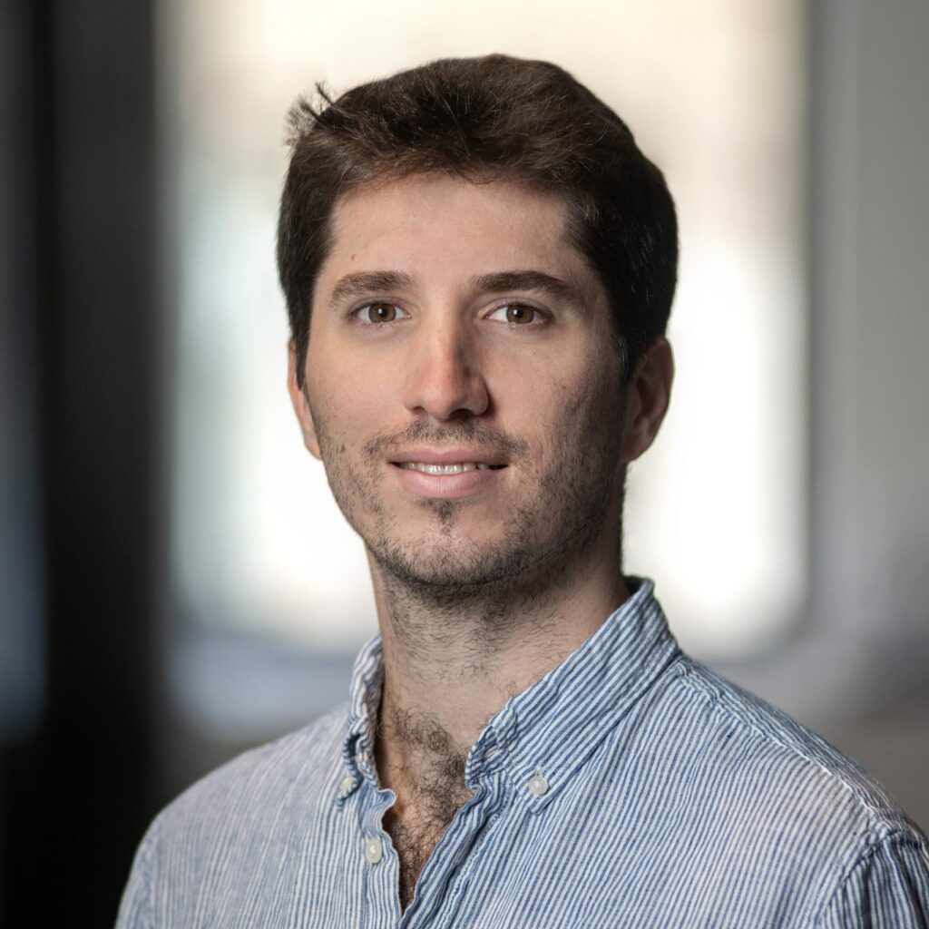 A young man with short brown hair and stubble is smiling slightly, wearing a light blue striped shirt. The background is softly blurred, highlighting the face.
