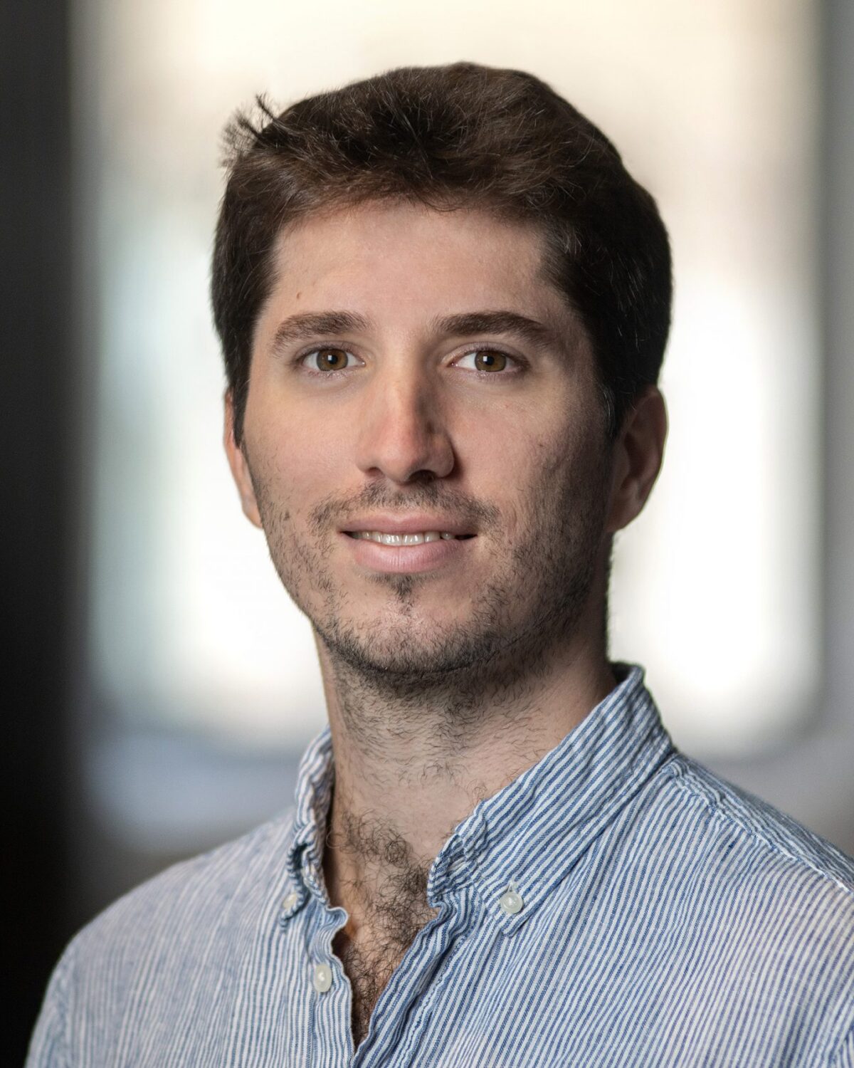 A young man with short brown hair and stubble is smiling slightly, wearing a light blue striped shirt. The background is softly blurred, highlighting the face.