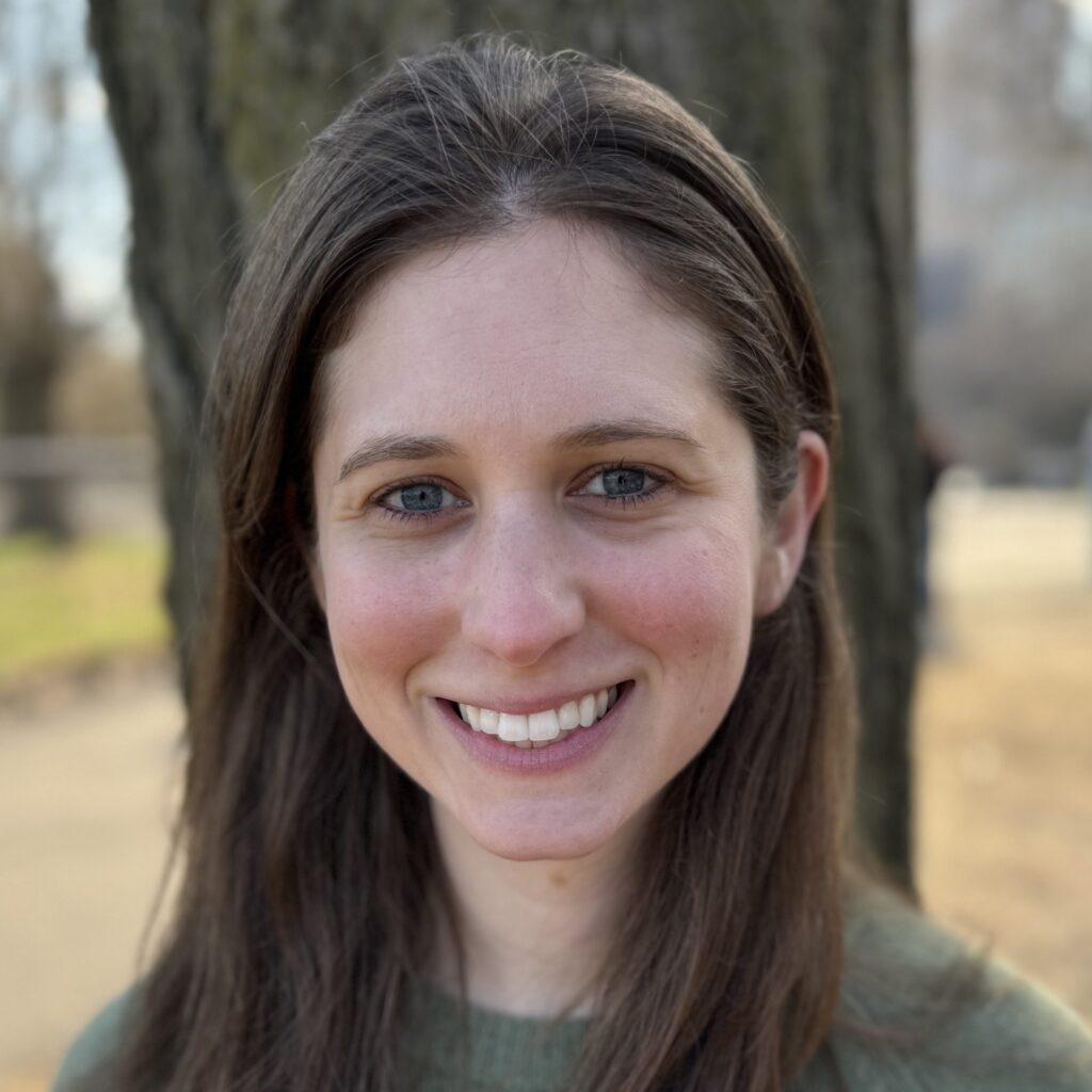 A woman with long brown hair and blue eyes smiles at the camera, standing outside in front of a tree on a sunny day.