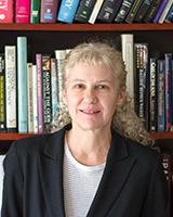 A woman with curly blonde hair wearing a black blazer and white shirt stands in front of a bookshelf filled with various books.