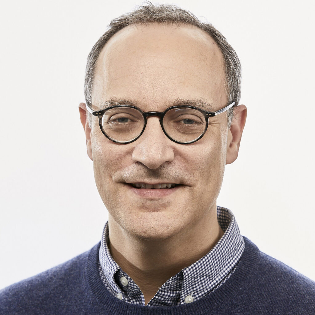 A man with short brown hair and glasses is wearing a blue sweater over a checkered shirt, posing against a plain white background.