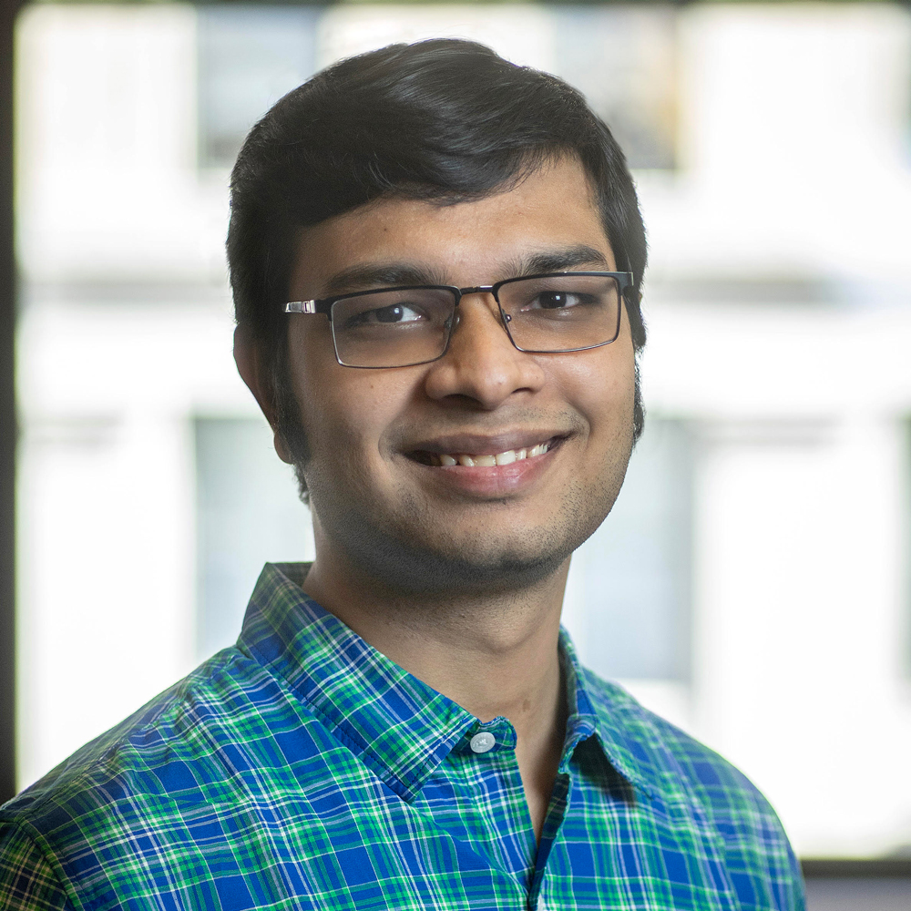 A man with short black hair and glasses smiles at the camera, wearing a blue and green plaid shirt, with a blurred bright background.