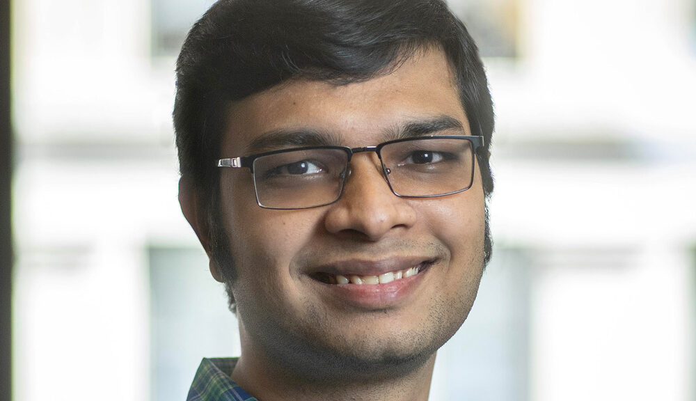 A man with short dark hair, glasses, and light facial hair is smiling at the camera. He is wearing a blue and green plaid shirt, and the background is softly blurred.