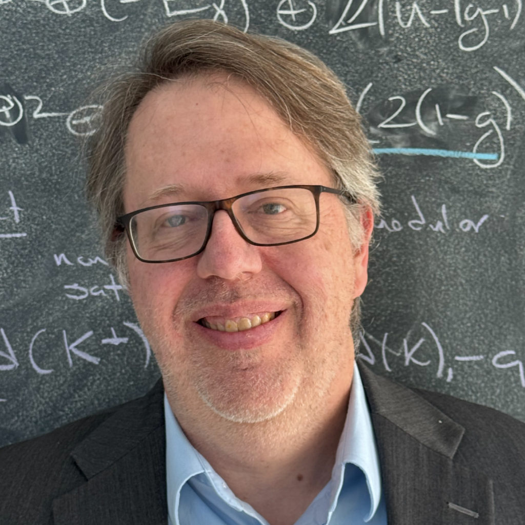 A man with glasses and graying hair smiles in front of a chalkboard filled with mathematical equations. He is wearing a blue shirt and a gray blazer.