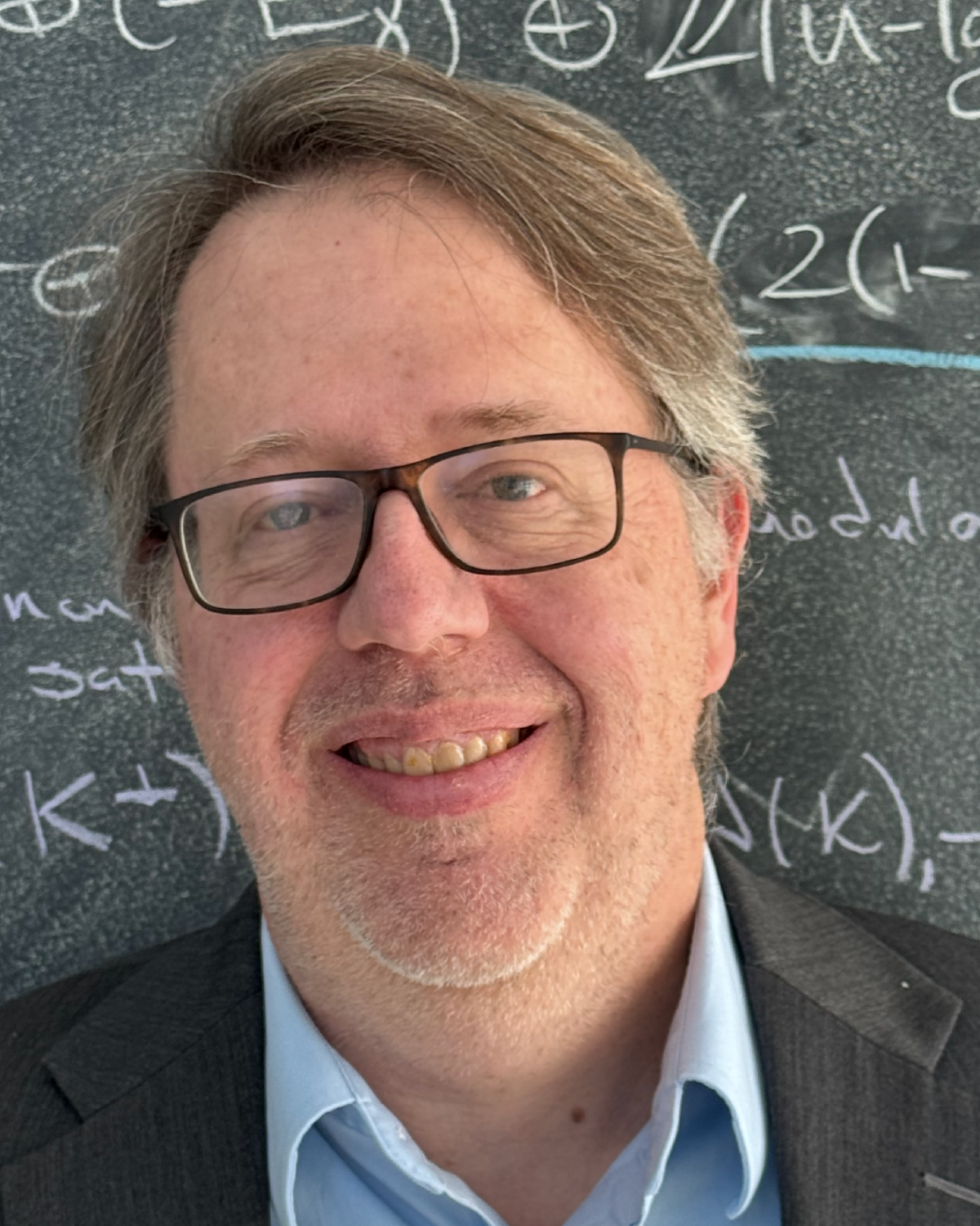 A man with glasses and graying hair smiles in front of a chalkboard filled with mathematical equations. He is wearing a blue shirt and a gray blazer.