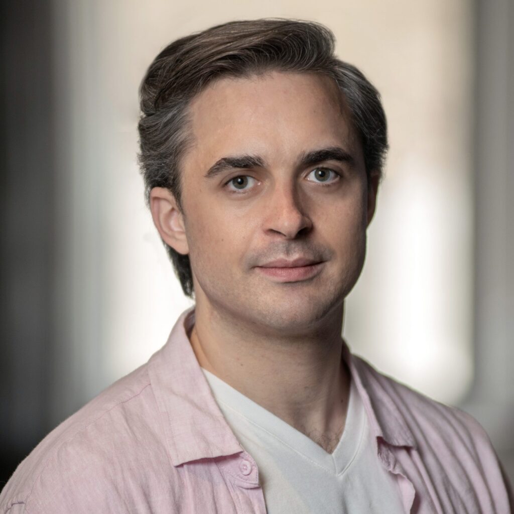 A person with short gray-brown hair wearing a light pink shirt over a white top looks at the camera with a neutral expression. The background is softly blurred.
