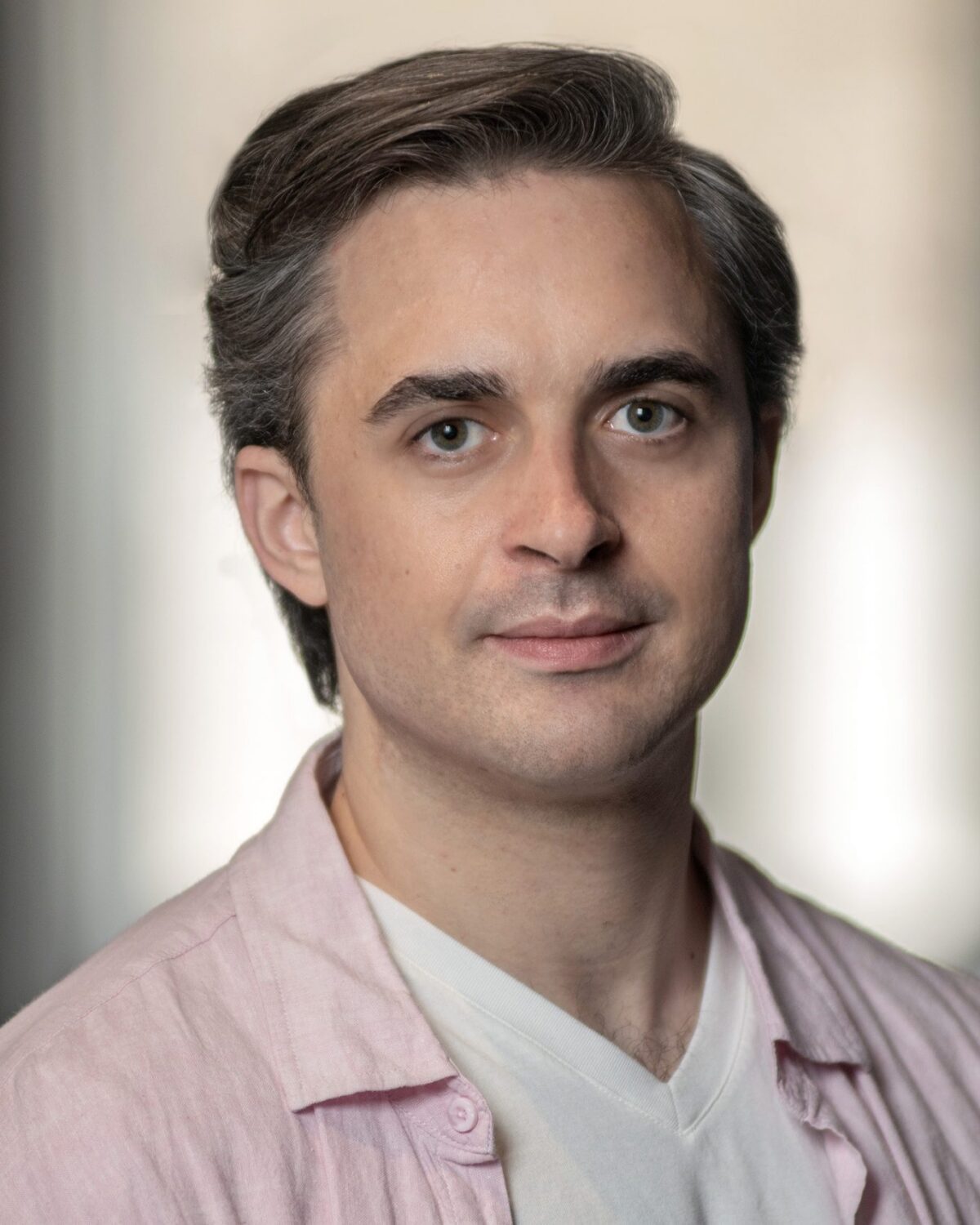 A person with short gray-brown hair wearing a light pink shirt over a white top looks at the camera with a neutral expression. The background is softly blurred.
