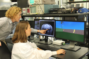Two people in a lab look at dual monitors; one shows a brain scan, the other a simple 3D scene with bananas. One person points at the brain scan while the other sits at the desk.