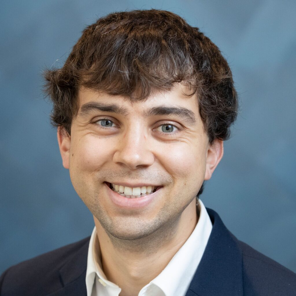 A smiling man with wavy brown hair wearing a dark suit jacket and white shirt, posing in front of a blue background.