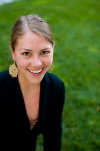 A young woman with light brown hair in a low bun, wearing large gold earrings and a dark top, smiles at the camera. She is outdoors, with green grass in the background.