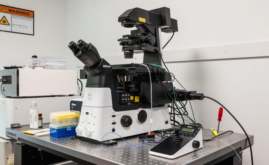 A Nikon laboratory microscope with attached cables and controls sits on a metal table, surrounded by lab equipment like pipette tips, bottles, and warning signs on the wall.