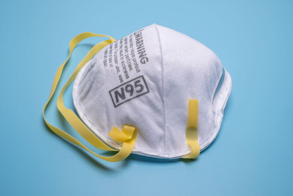 A white N95 respirator mask with yellow elastic bands rests on a light blue surface. The mask displays warning text and the N95 label in bold print on the front.