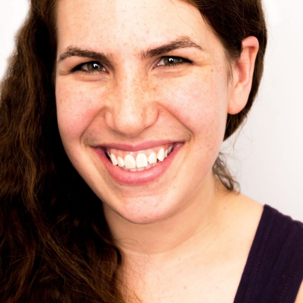 A woman with long brown hair and freckles, wearing a sleeveless dark top, smiles at the camera against a plain white background.