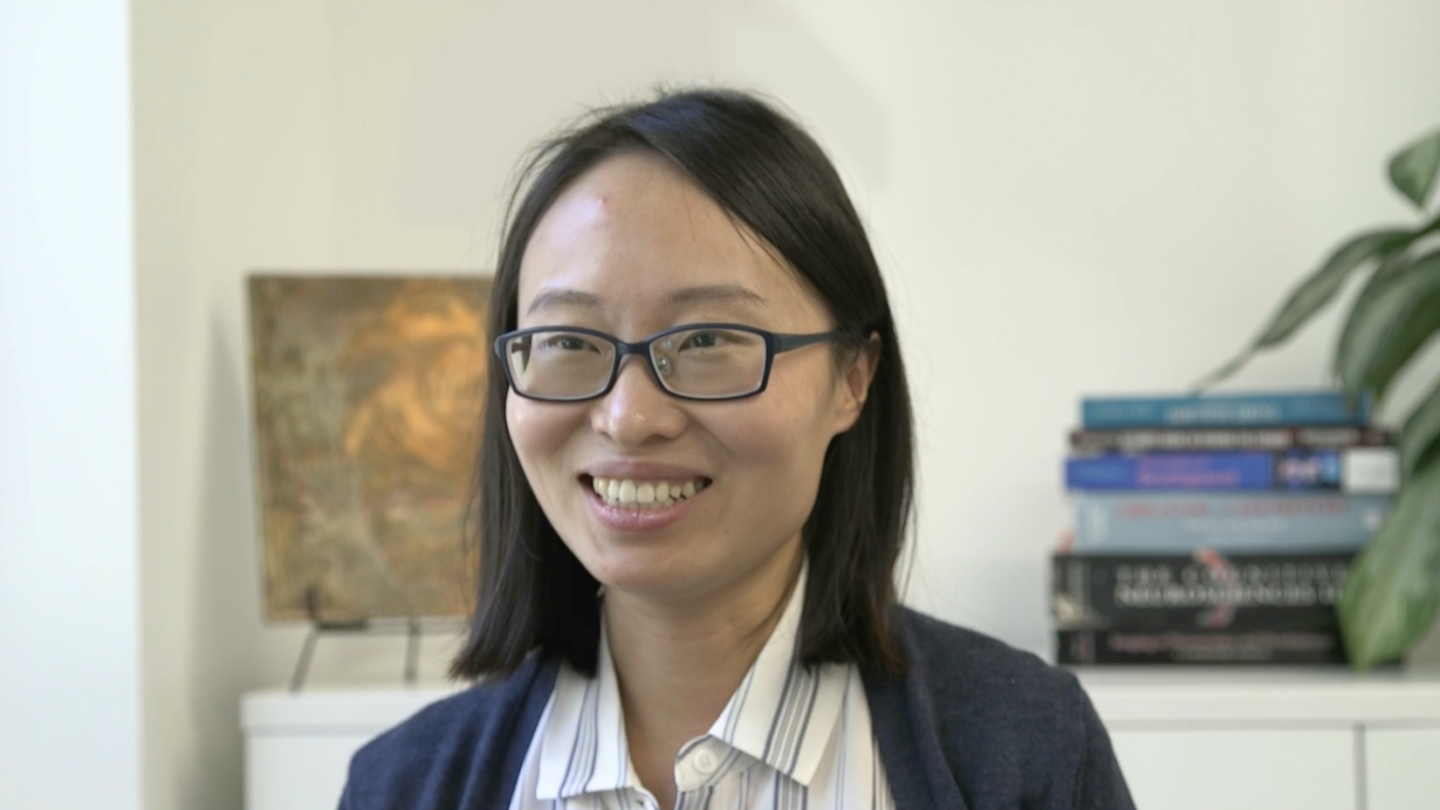 A woman with glasses and shoulder-length dark hair smiles in an indoor setting, with books and a plant visible in the background.