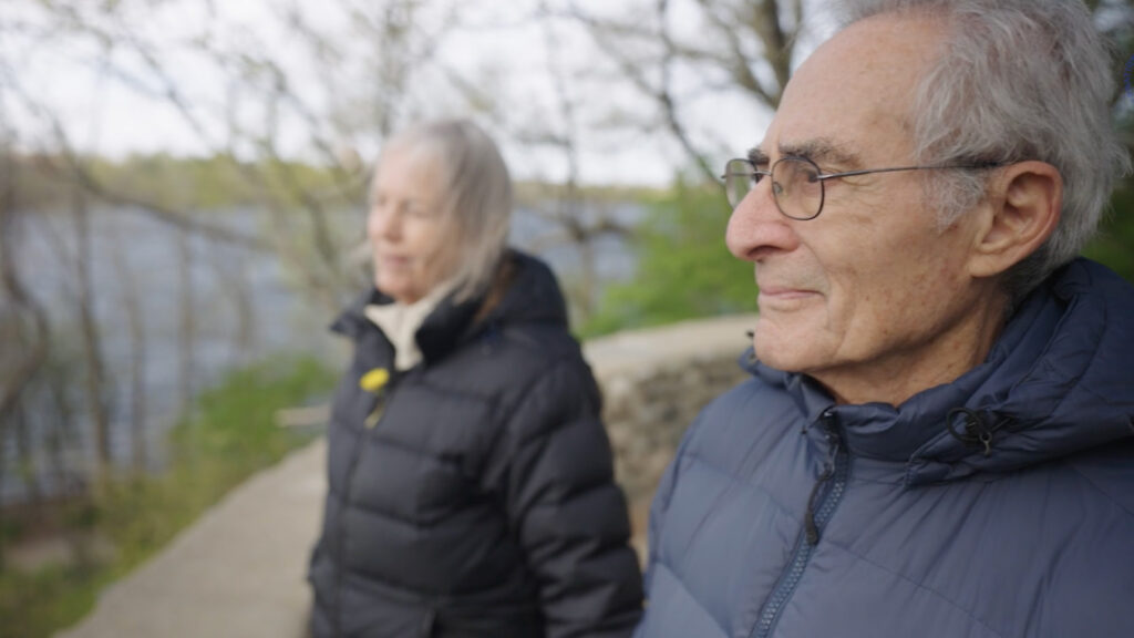 An older man and woman wearing dark jackets stand outdoors near water, looking off into the distance with trees and greenery in the background.