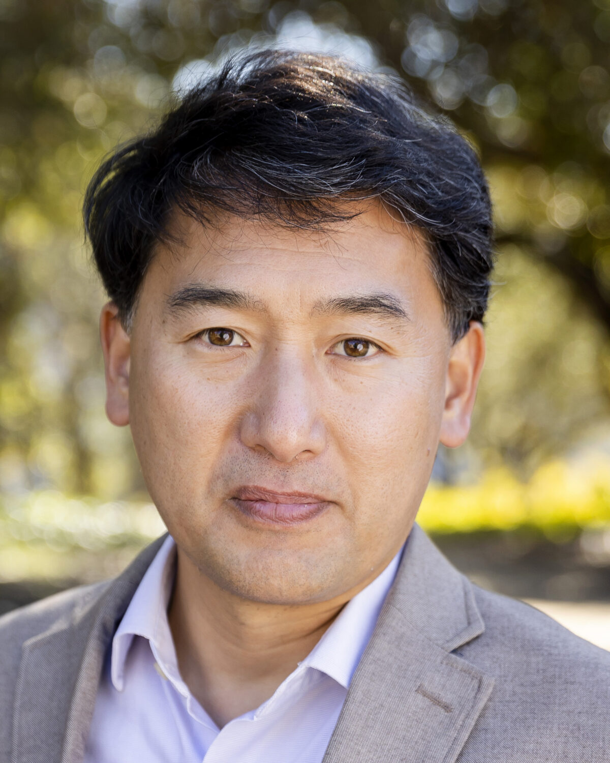 A man with short dark hair wearing a beige blazer and light shirt stands outdoors, with trees and sunlight blurred in the background.