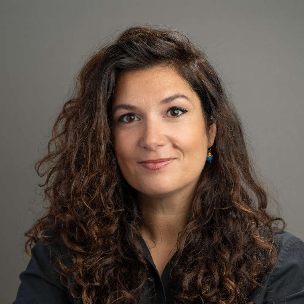 Woman with long curly brown hair, wearing a black shirt and blue earrings, poses in front of a plain gray background, smiling softly at the camera.