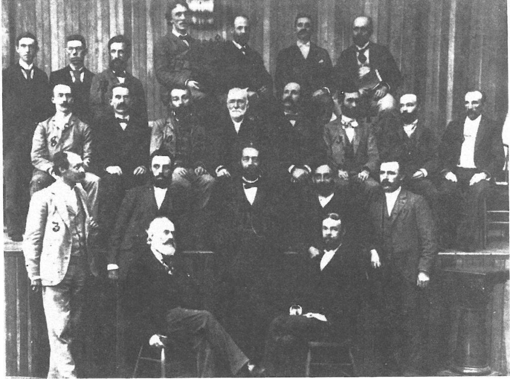 A group of about twenty men in suits and ties pose together indoors, some seated and others standing, against a striped wall. The image is in black and white and has a vintage, historical appearance.