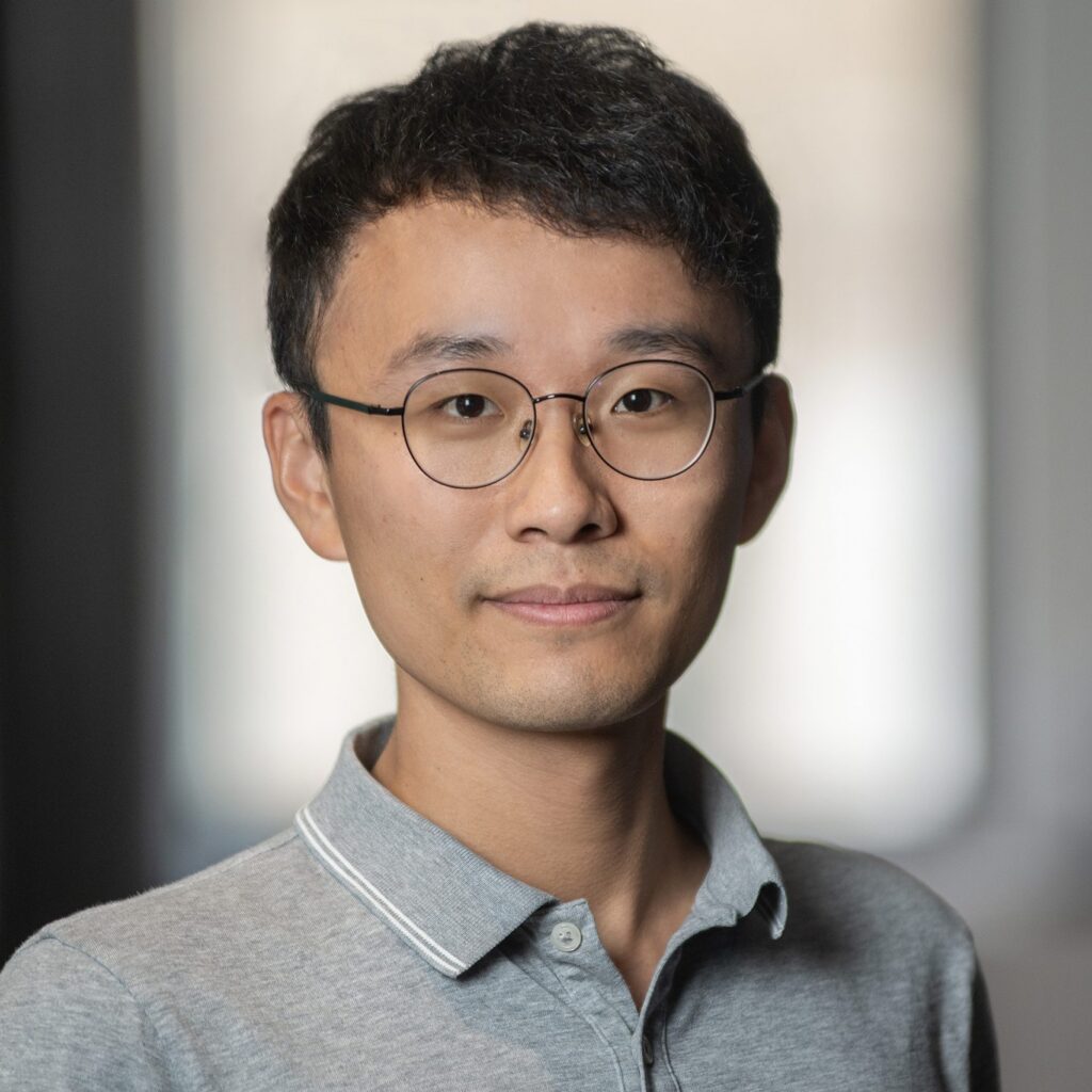 A young person with short black hair and glasses, wearing a gray collared shirt, stands indoors in front of a blurred background, looking at the camera with a neutral expression.