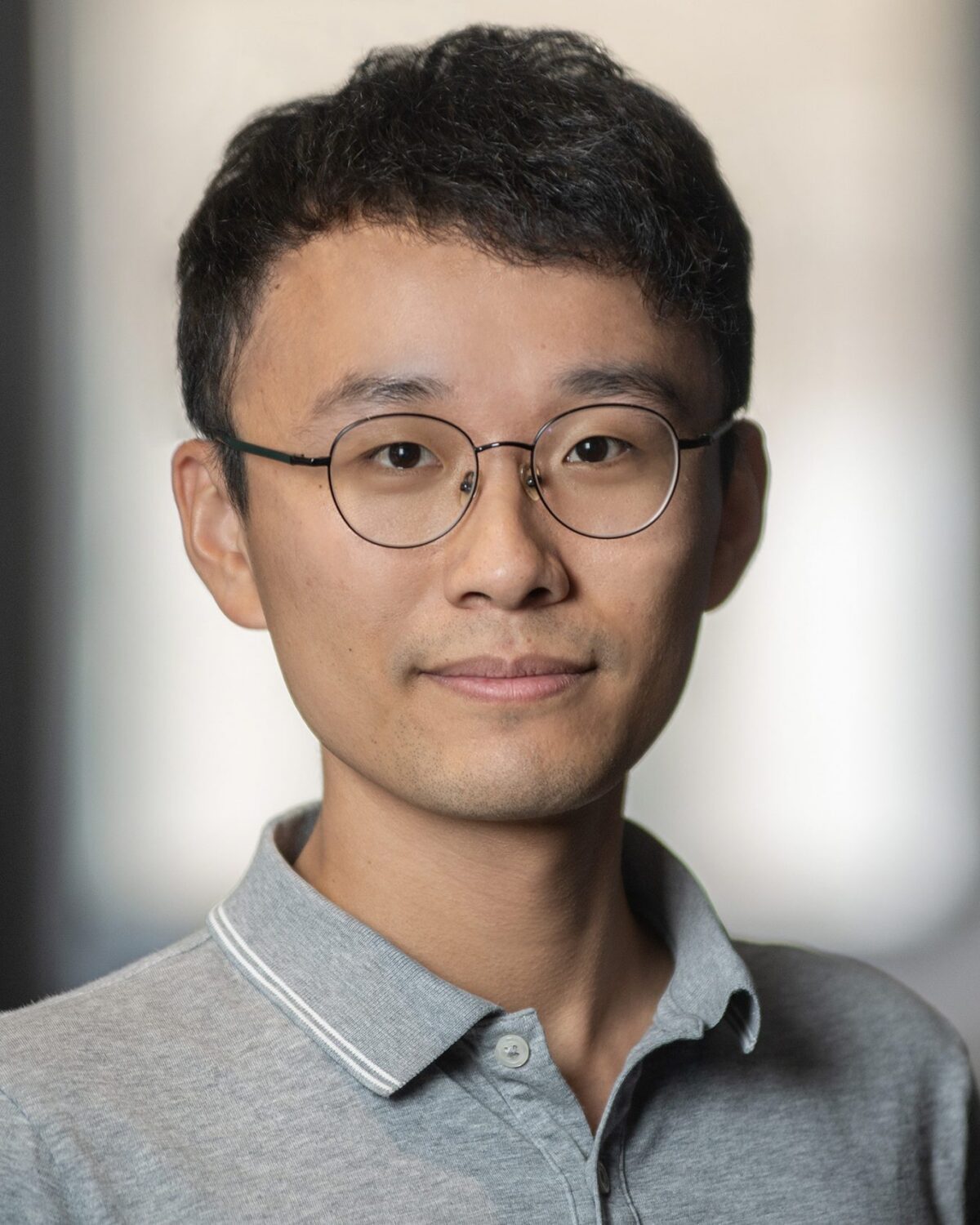 A young person with short black hair and glasses, wearing a gray collared shirt, stands indoors in front of a blurred background, looking at the camera with a neutral expression.