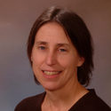 Woman with straight brown hair, wearing a dark top, smiling at the camera against a neutral, brownish background.
