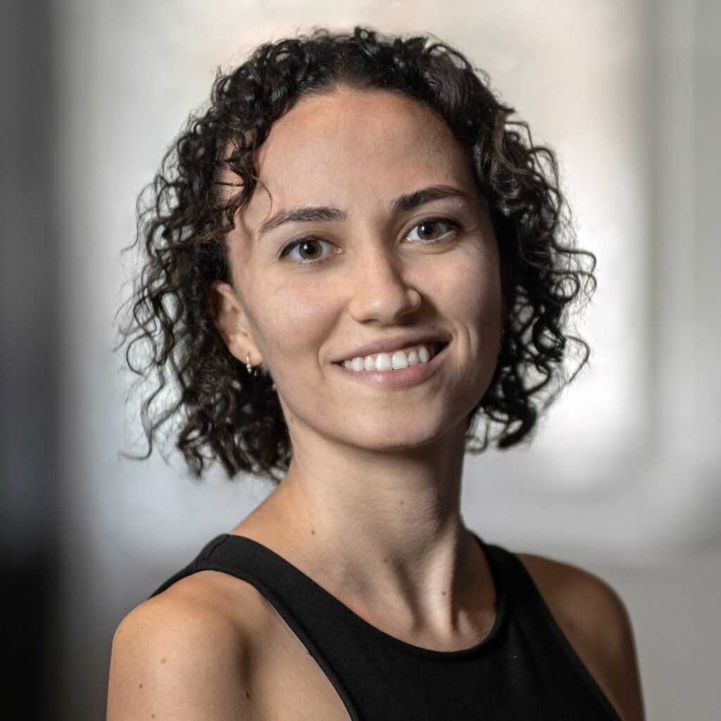 A woman with curly dark hair, wearing a sleeveless black top, smiles at the camera in a softly blurred indoor setting.