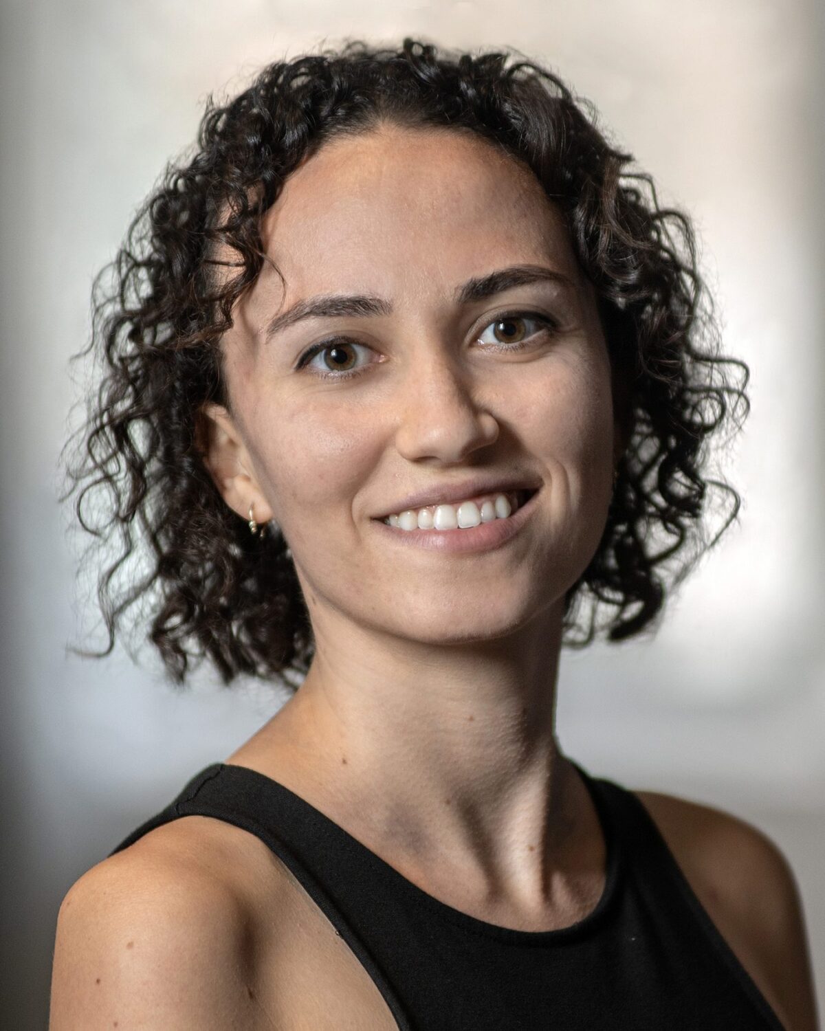 A woman with curly dark hair, wearing a sleeveless black top, smiles at the camera in a softly blurred indoor setting.